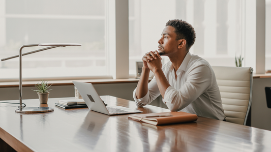 A young man sitting at a corporate desk thinking in front of his laptop.
